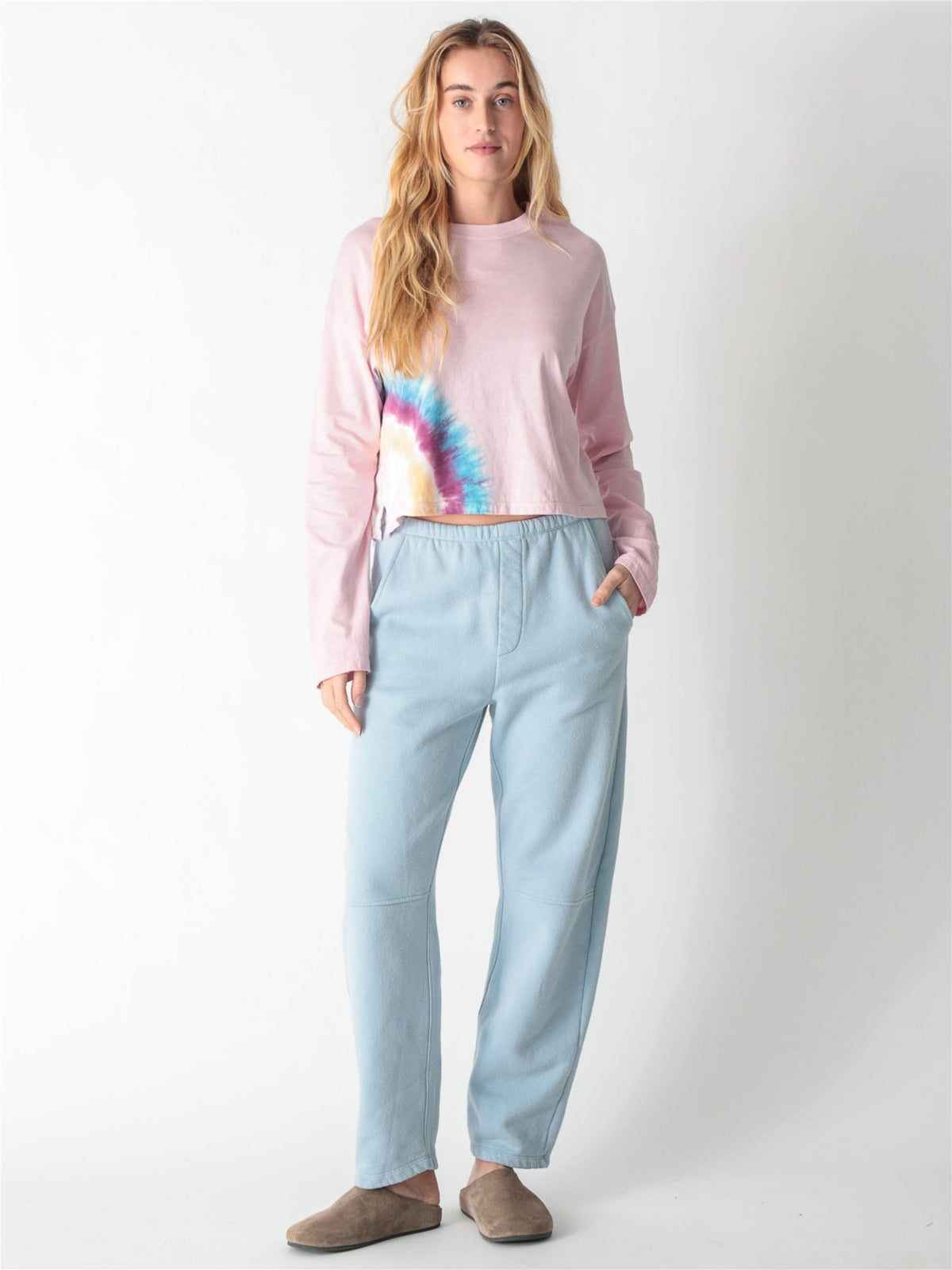 A woman wearing a light pink long-sleeve crop top featuring a colorful tie-dye accent along the right hem, paired with light blue sweatpants against a white background.