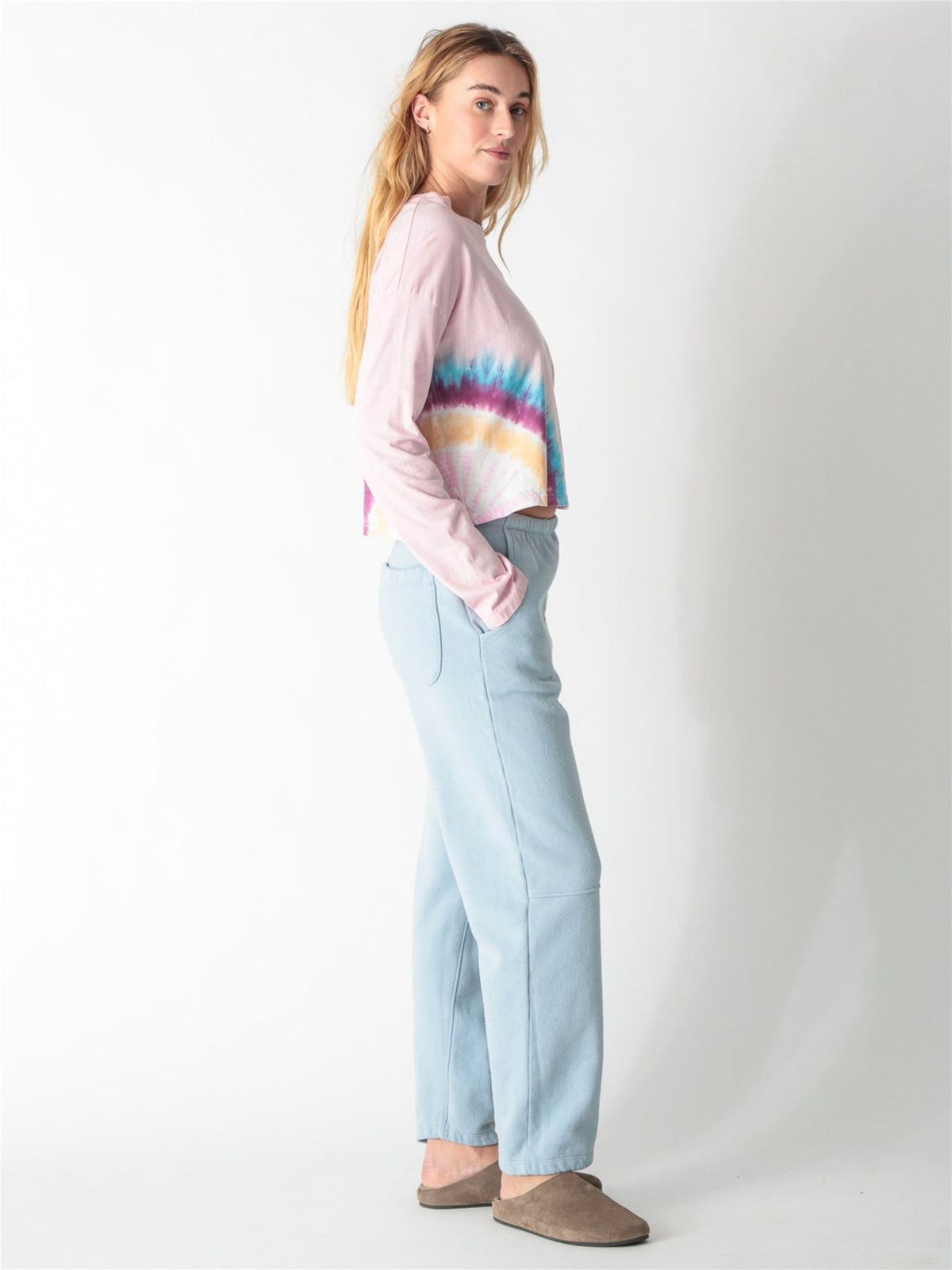 A woman wearing a light pink long-sleeve crop top featuring a colorful tie-dye accent along the right hem, paired with light blue sweatpants against a white background.
