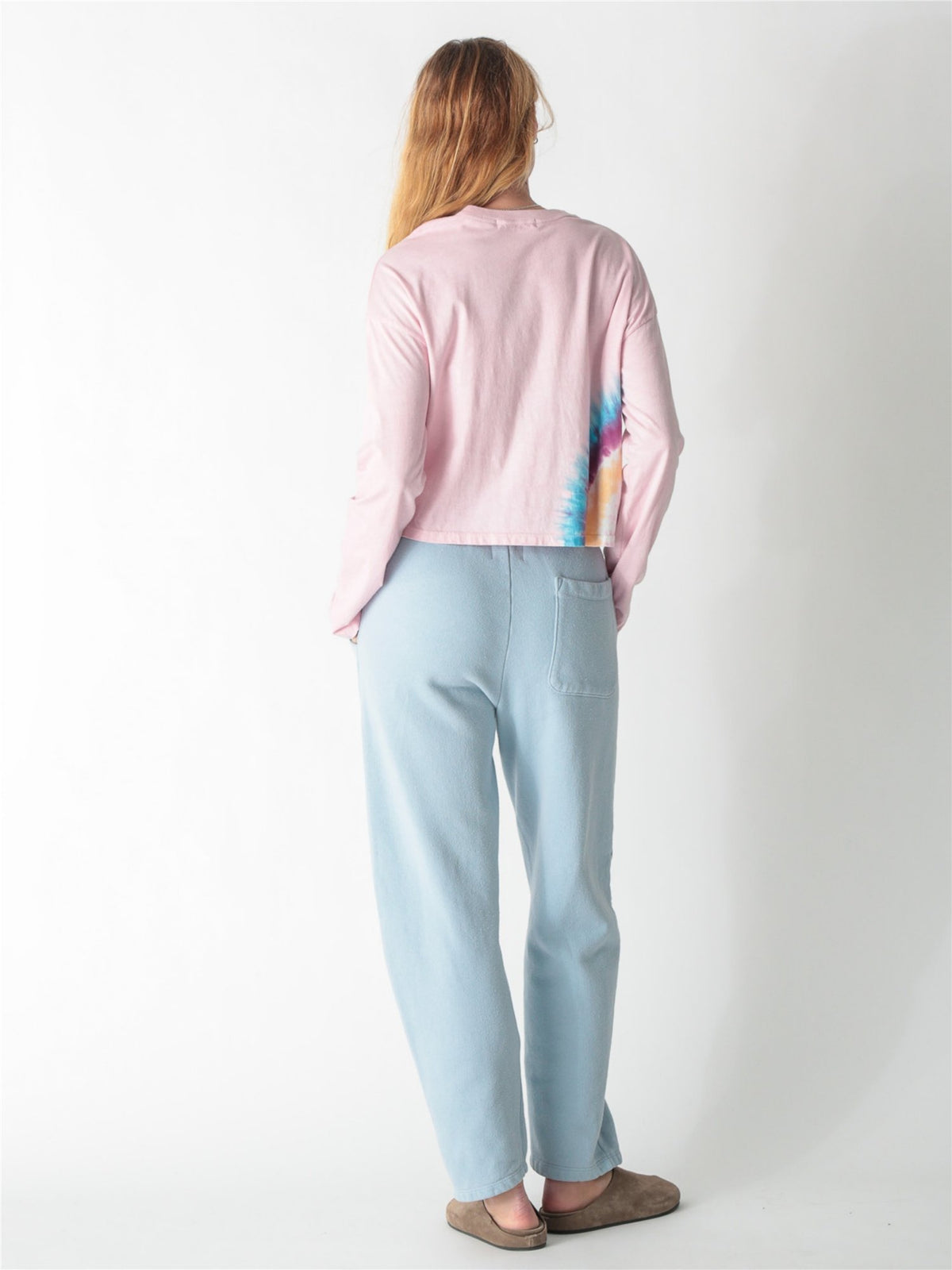 A woman wearing a light pink long-sleeve crop top featuring a colorful tie-dye accent along the right hem, paired with light blue sweatpants against a white background.