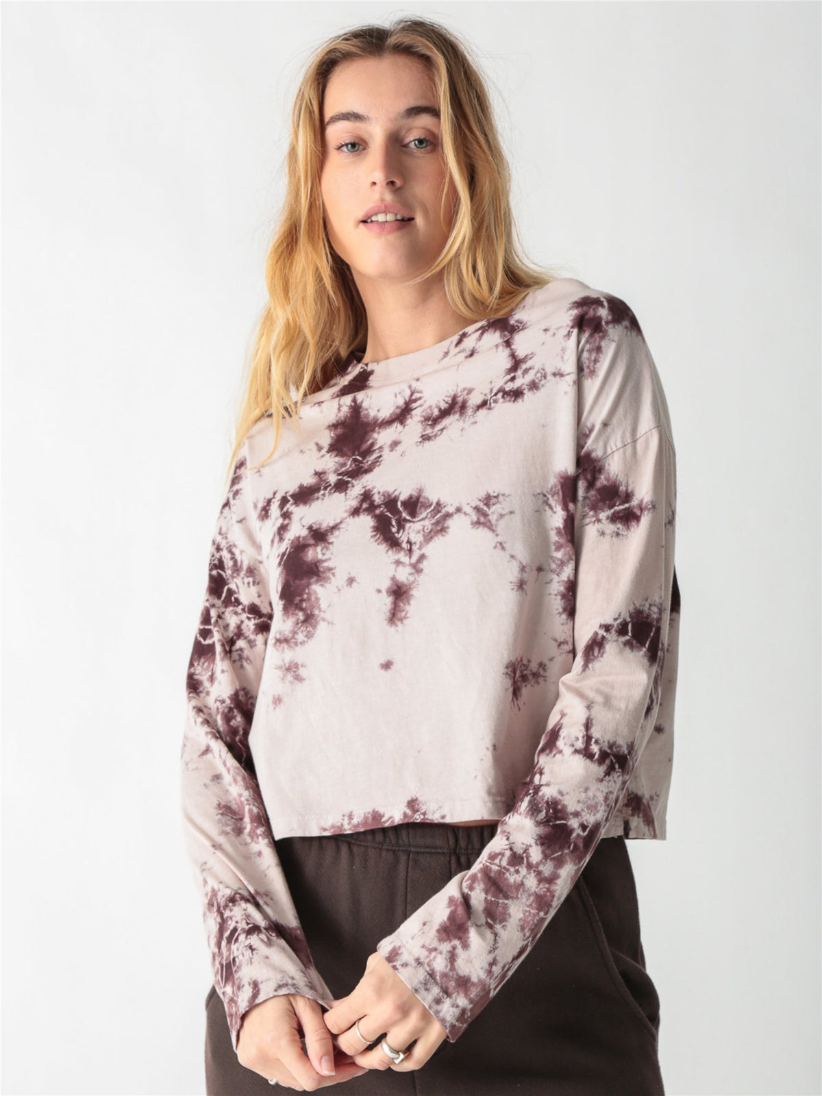 A woman wearing a long-sleeve cropped t-shirt with a cream and brown tie-dye pattern, paired with dark brown pants against a white background.