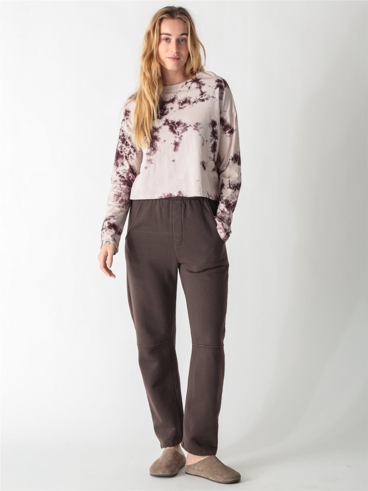 A woman wearing a long-sleeve cropped t-shirt with a cream and brown tie-dye pattern, paired with dark brown pants against a white background.