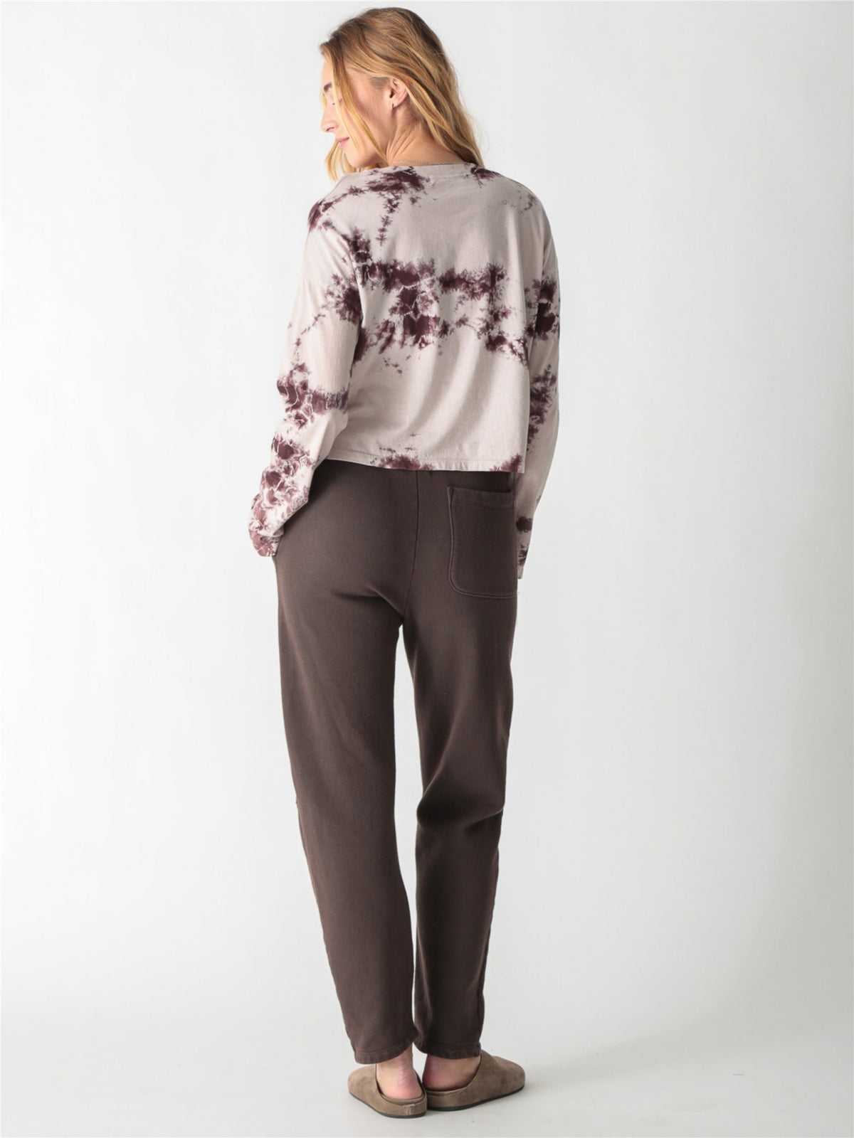 A woman wearing a long-sleeve cropped t-shirt with a cream and brown tie-dye pattern, paired with dark brown pants against a white background.